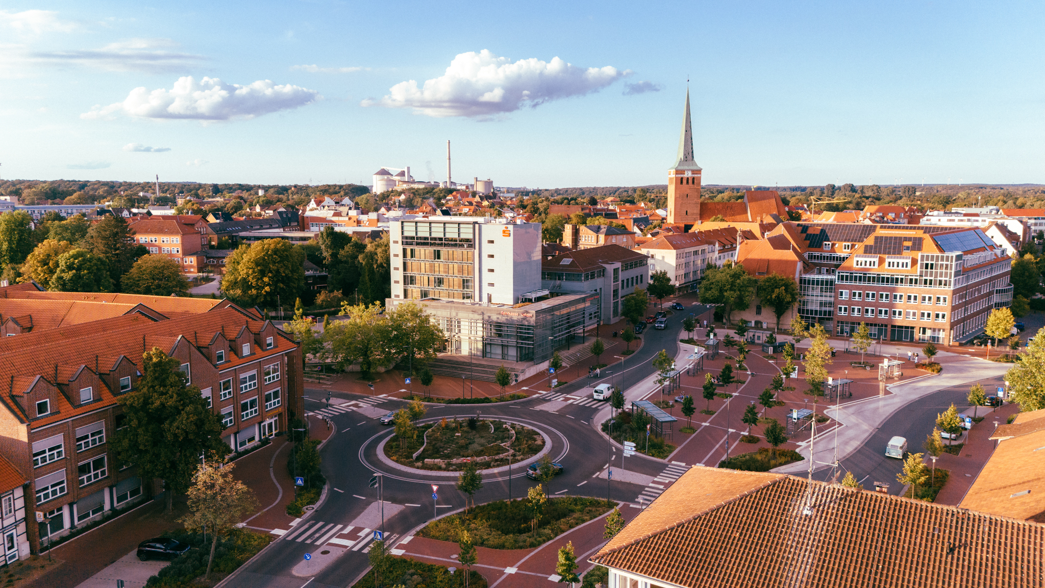 Luftaufnahme der Uelzener Skyline mit großem Kreisverkehr im Fokus neben vielen Häusern & Kirche zwischen einigen Bäumen, im Hintergrund ein hellblauer Horizont.