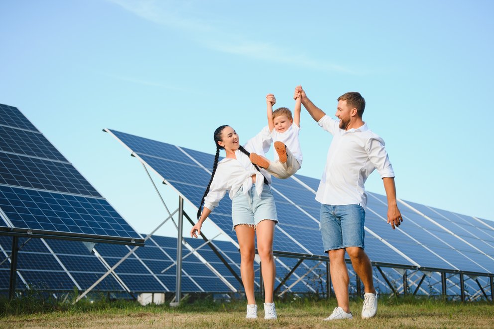 Ein Frau und ein Mann heben ihr Kind zwischen Ihnen an den Armen hoch und lassen es in der Luft fliegen. Im Hintergrund ein Solarpark.